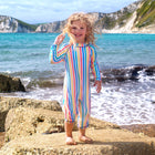 A child wearing a UV Protective Surf Suit Multi Stripe stands on rocks at the beach with the sea and cliffs in the background.